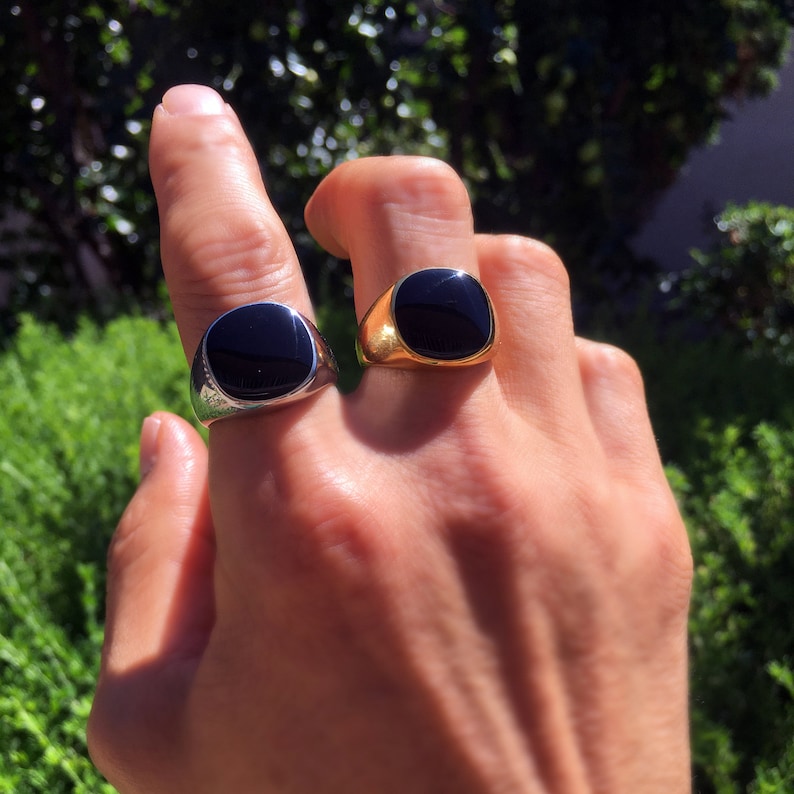 May include: Two signet rings featuring smooth, polished black stone inlays. One ring is crafted from a bright silver-toned metal, while the other is made from a warm gold-toned metal. Both rings have a rounded square face and a solid band design.