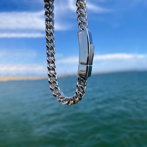 May include: A silver-toned curb chain necklace featuring interlocking polished metal links and a rectangular box clasp closure. The chain is suspended vertically, showcasing the reflective surface of the metal links and the secure locking mechanism.