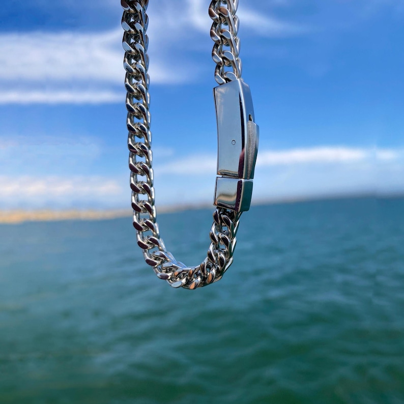 May include: A silver-toned curb chain necklace featuring interlocking polished metal links and a rectangular box clasp closure. The chain is suspended vertically, showcasing the reflective surface of the metal links and the secure locking mechanism.