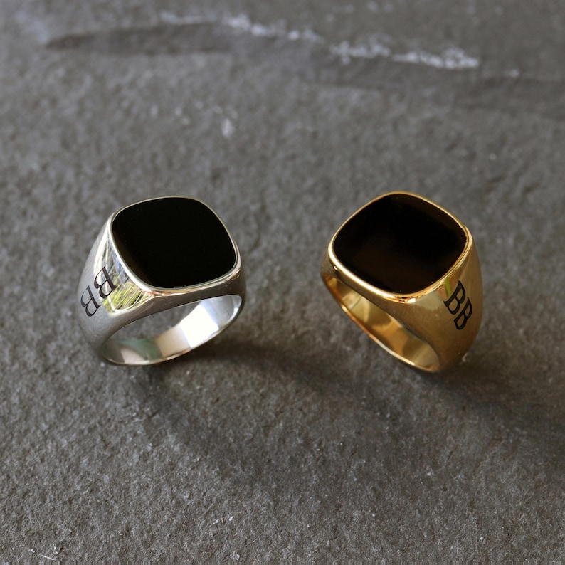 May include: Two signet rings featuring a square face with rounded corners and a smooth black inlay. One ring is polished silver-tone metal and the other is polished gold-tone metal. Both rings have the letters BB engraved on the side of the band.