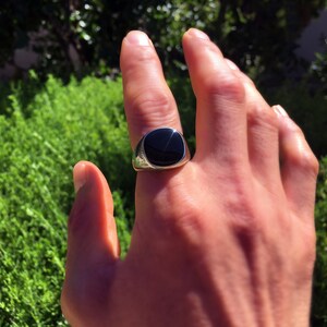May include: A silver signet ring featuring a smooth, polished black stone inlay. The ring has a rounded, metallic band that tapers toward the back and a flat, circular face that holds the dark, reflective stone securely in the center setting.