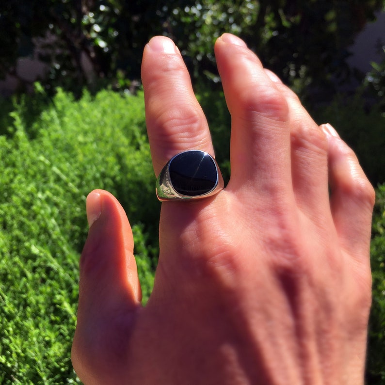 May include: A silver signet ring featuring a smooth, polished black stone inlay. The ring has a rounded, metallic band that tapers toward the back and a flat, circular face that holds the dark, reflective stone securely in the center setting.