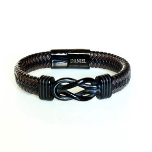 May include: A black leather bracelet with a brown leather braid and a black metal clasp. The clasp has the word "DANIEL" engraved on it.