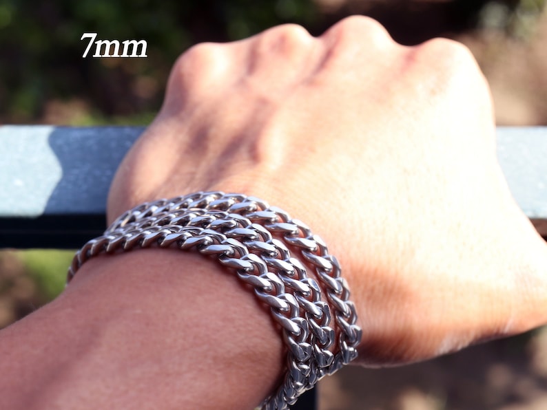 K&ouml;nnte beinhalten: Ein silbernes Kettenarmband mit einer Breite von 7 mm. Das Armband ist an einem Handgelenk &uuml;bereinander gelegt.