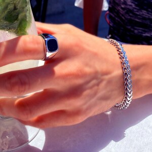 May include: A silver-toned signet ring featuring a smooth, rectangular black inlay and a silver-toned chain link bracelet with a woven, interlocking pattern. Both pieces of jewelry are polished and have a metallic, reflective finish.