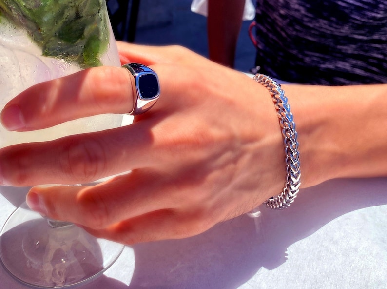 May include: A silver-toned signet ring featuring a smooth, rectangular black inlay and a silver-toned chain link bracelet with a woven, interlocking pattern. Both pieces of jewelry are polished and have a metallic, reflective finish.