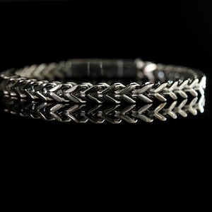 May include: A silver-toned metal bracelet featuring a repeating chevron or wheat-style chain link pattern. The bracelet has a polished, reflective surface and a rectangular clasp mechanism visible at the center back of the circular band.