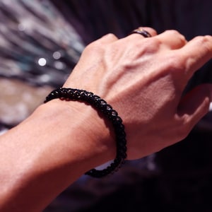 May include: A black chain link bracelet with a polished finish worn on the wrist. The bracelet features a series of interconnected oval links that form a continuous band around the arm, showcasing a dark, reflective metallic surface texture.
