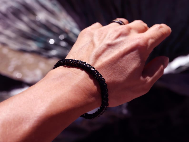 May include: A black chain link bracelet with a polished finish worn on the wrist. The bracelet features a series of interconnected oval links that form a continuous band around the arm, showcasing a dark, reflective metallic surface texture.