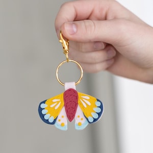 May include: A colorful butterfly keychain with a gold-colored clasp and ring. The butterfly has yellow wings with blue and white accents, a red body, and white spots. The keychain is held by a hand.