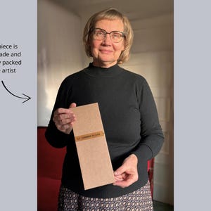 Professional textile artist Elina Lūsis-Grīnberga holding a carefully packaged handmade artwork, labeled “ELG | Handmade in Latvia” – each piece is individually crafted and packed with care.