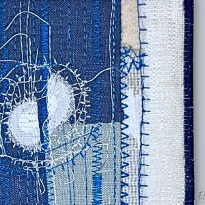 Close-up of indigo embroidered textile art showing fine stitching and layered fabric textures in white, navy, and linen tones.