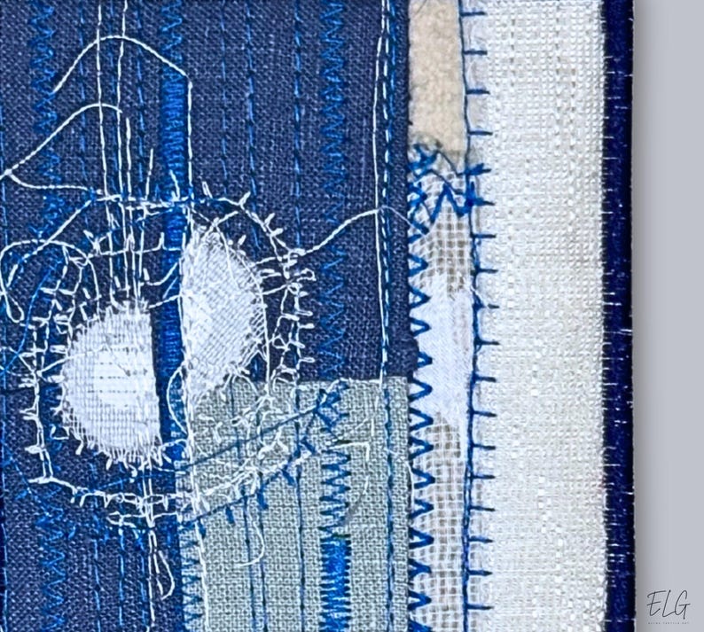 Close-up of indigo embroidered textile art showing fine stitching and layered fabric textures in white, navy, and linen tones.