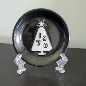 May include: A round black ceramic plate featuring a white embossed Christmas tree design in the center. The tree is topped with a star and contains the numbers 1 9 7 6 arranged vertically. The plate is displayed on a clear plastic stand.
