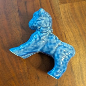 May include: A blue ceramic figurine of a dog with a bow around its neck. The dog is in a sitting position with its front paws extended. The figurine has a glossy finish and is a medium shade of blue.