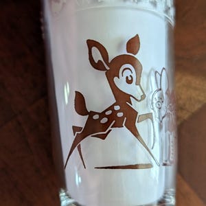 May include: Clear glass tumbler featuring a brown Bambi graphic and a rabbit. The top rim has a decorative band with animal silhouettes. A classic design for fans of vintage glassware.