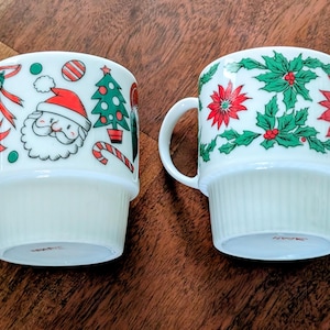 May include: Two white ceramic Christmas mugs. One mug features Santa Claus, a Christmas tree, and candy cane designs. The other mug has a pattern of red poinsettias and green holly leaves. Both mugs have a handle.