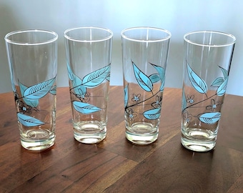 May include: A set of four clear glass tumblers, each featuring a decorative wrap-around design of light blue leaves and small black-outlined flowers. The tall, cylindrical drinking glasses have a smooth finish and a weighted base for stability.