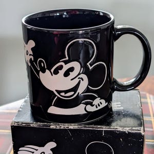 May include: Black ceramic mug featuring a white embossed image of Mickey Mouse waving. The mug has a rounded handle and is sitting on a black box with a matching Mickey Mouse design. The mug is a collectible item.