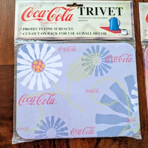 May include: A Coca-Cola trivet with a floral design and Coca-Cola branding. The trivet is packaged and features the text "PROTECTS FINE SURFACES" and "CUT-OUT ON BACK FOR USE AS WALL DECOR". The trivet is a nostalgic collectible.
