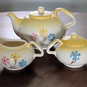 May include: A vintage tea set featuring a teapot, creamer, and sugar bowl. The set is cream-colored with yellow accents and floral designs in pink, yellow, and blue. The teapot has a lid and a curved handle. The sugar bowl also has a lid.