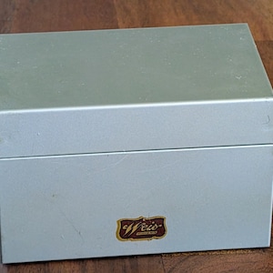 May include: A silver-colored metal box with a hinged lid. The box has a rectangular shape and a smooth surface. A small, ornate label with the word "Weis" is on the front of the box.