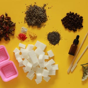 Puede incluir: Vista aérea de suministros para hacer jabón sobre una superficie amarilla. Incluye flores secas, hierbas, granos de café, semillas de chía, una botella de aceite esencial, moldes de jabón y cubos de base de jabón. Los moldes rosas dicen "100% HAND MADE".