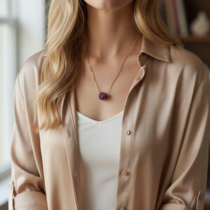 Könnte beinhalten: Eine goldene Halskette mit einem rohen, dunkelvioletten Amethyst-Anhänger. Die Halskette wird über einem weißen Oberteil und einem beigefarbenen Hemd getragen.