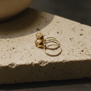 Könnte beinhalten: Ein Paar goldene Creolen mit tropfenförmigen, facettierten Edelsteinen. Die Ohrringe werden auf einer strukturierten, hellfarbigen Oberfläche präsentiert. Die Creolen sind dünn und filigran und reflektieren das Licht.