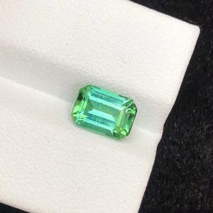 Op de afbeelding: Een rechthoekige, smaragdgeslepen edelsteen met een levendige groene kleur. De edelsteen ligt op een wit oppervlak, waardoor de helderheid en schittering worden benadrukt. De facetten van de steen reflecteren het licht, waardoor een sprankelend effect ontstaat. Ideaal voor het maken van sieraden.