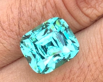May include: A vibrant, rectangular-cut gemstone with a striking turquoise hue. The gem displays multiple facets, creating a brilliant display of light and color. The stone is held in a hand, showcasing its size and clarity.