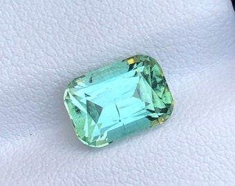 May include: A rectangular-cut gemstone with a light teal color and a faceted surface. The gem has a slight green hue and is set against a white background. The gemstone is likely a precious stone, such as a tourmaline or aquamarine.