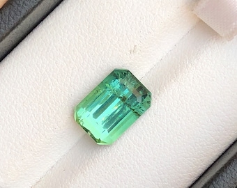 May include: A rectangular, faceted, teal-green gemstone rests in a black jewelry box with a white interior. The gem has a gradient of color, transitioning from a darker green at the top to a lighter, almost transparent green at the bottom. The gem is cut with multiple facets.