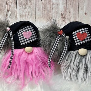 May include: Two gnomes with black hats and white and black checkered fabric accents. One gnome has pink fur and a pink heart on its hat, the other has gray fur and a red heart on its hat.