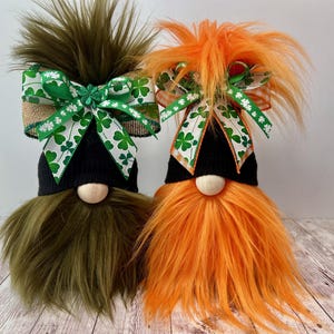 May include: Two St. Patrick's Day gnomes with black hats, wooden noses, and faux fur beards. One gnome has a green beard and the other has an orange beard. Both have bows with shamrock patterns.