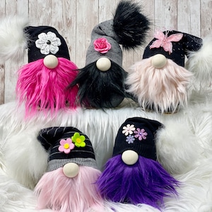 May include: Six gnomes with different colored fur and hats decorated with flowers and rhinestones. The gnomes are sitting on a white fluffy surface.