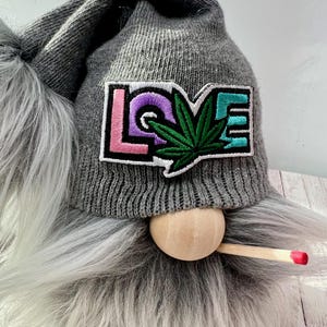 May include: A gnome with a gray knit cap and long gray beard. The cap features a patch with the word "LOVE" and a green cannabis leaf. A wooden nose and a small, red-tipped wooden object complete the design.