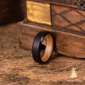 Puede incluir: Un anillo negro con exterior de metal cepillado e interior de madera clara. El anillo se encuentra sobre un fondo de caja de madera y mapa vintage. El dise&ntilde;o del anillo es simple y moderno.