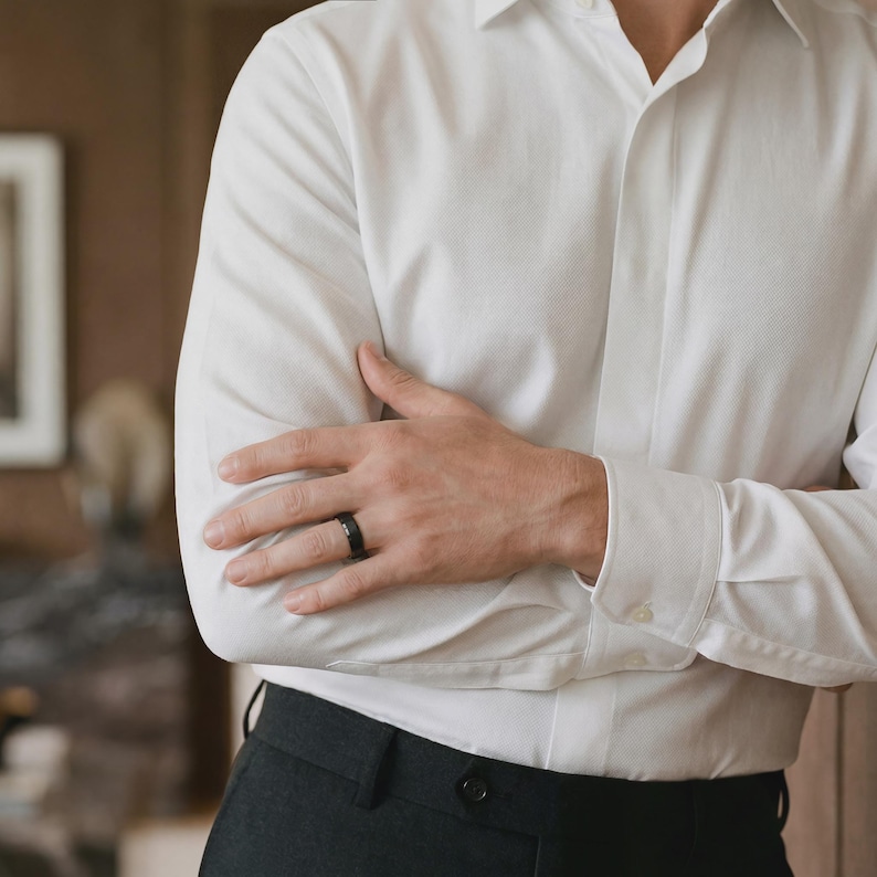 Puede incluir: Un anillo negro en el dedo de una persona. El anillo se lleva con una camisa blanca y pantalones grises oscuros. Los brazos de la persona est&aacute;n cruzados. El fondo est&aacute; borroso.