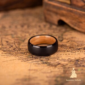Puede incluir: Un anillo negro con interior de madera clara descansa sobre un mapa antiguo. El anillo tiene un acabado mate y una banda ancha. El interior del anillo es de color marr&oacute;n claro.