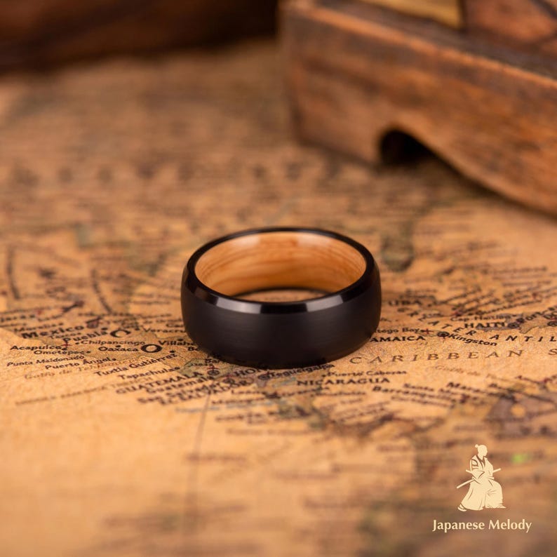 Puede incluir: Un anillo negro con interior de madera clara descansa sobre un mapa antiguo. El anillo tiene un acabado mate y una banda ancha. El interior del anillo es de color marr&oacute;n claro.