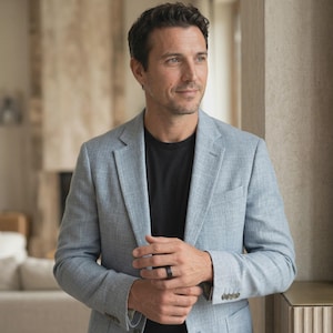 Puede incluir: Un blazer azul claro sobre una camiseta negra. El blazer tiene un dise&ntilde;o cl&aacute;sico. Se ve un anillo negro en la mano izquierda. La persona est&aacute; de pie cerca de una ventana.