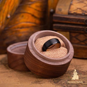 Puede incluir: Un anillo negro con interior de madera descansa en una caja de anillos de madera abierta, forrada con arpillera. Otra caja es visible en el fondo. El anillo es el punto focal.