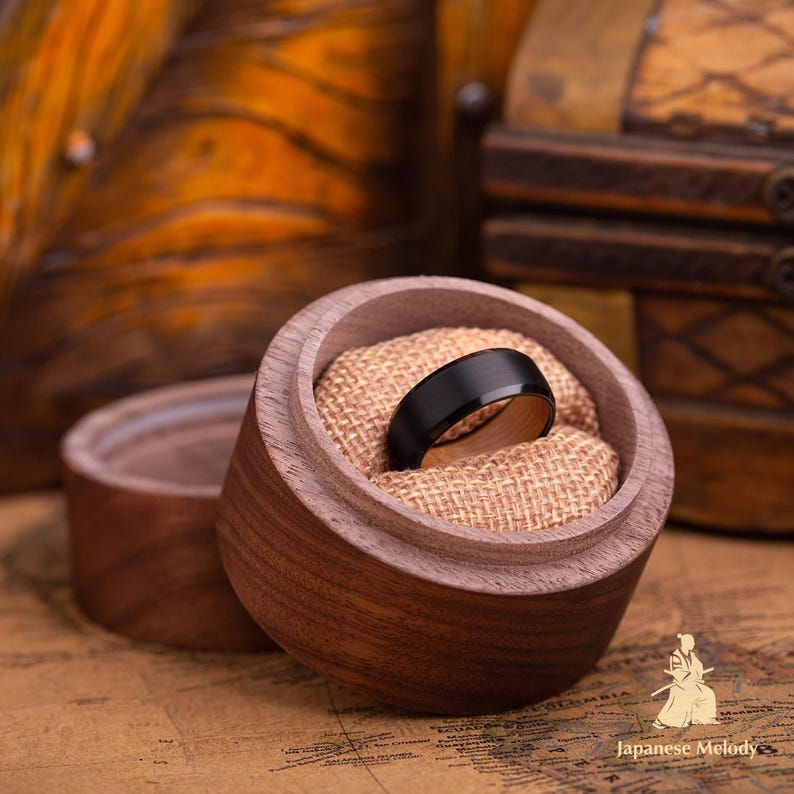 Puede incluir: Un anillo negro con interior de madera descansa en una caja de anillos de madera abierta, forrada con arpillera. Otra caja es visible en el fondo. El anillo es el punto focal.