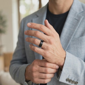 Puede incluir: Un anillo negro con acabado cepillado se lleva en un dedo. El anillo tiene un dise&ntilde;o simple y moderno. La persona lleva una blazer azul claro y una camisa azul oscuro.