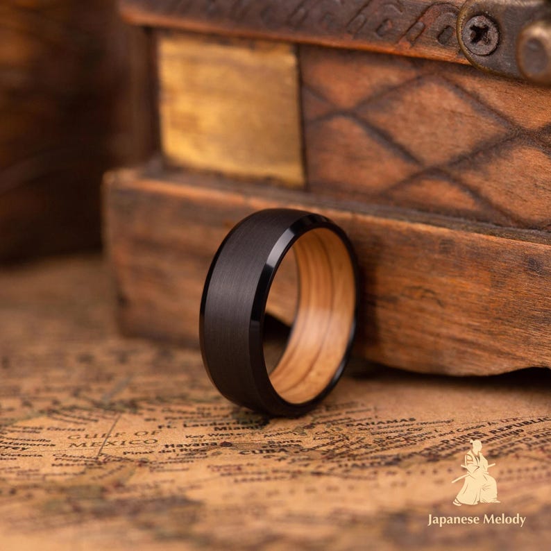 Puede incluir: Un anillo negro con interior de madera. El anillo tiene un acabado oscuro y mate en el exterior y un interior de veta de madera marr&oacute;n claro. El anillo es de estilo banda, con una superficie lisa y redondeada. El anillo est&aacute; sobre un mapa.