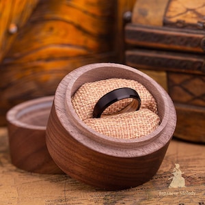 Puede incluir: Un anillo negro con acabado pulido descansa dentro de una caja redonda de madera forrada con arpillera. La caja est&aacute; abierta, revelando el dise&ntilde;o elegante del anillo. El anillo se exhibe sobre un mapa, sugiriendo un tema de viaje o aventura.