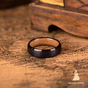 Puede incluir: Un anillo negro con interior de madera descansa sobre un mapa. El anillo tiene un exterior negro mate y un interior de veta de madera marr&oacute;n claro. El anillo es de dise&ntilde;o sencillo.