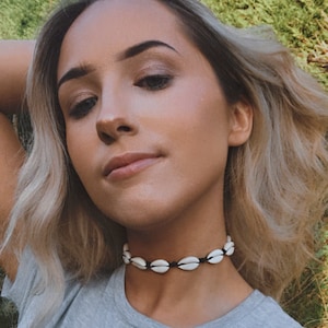 May include: A choker necklace made of white seashells on a black cord. The shells are oval and evenly spaced. The necklace is worn against a grey t-shirt.