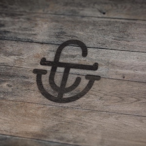May include: A dark, stylized monogram logo is cast as a shadow on a weathered, wooden surface. The logo features a stylized "f" intertwined with a "t" and a curved element, creating a unique design. The wood grain adds texture.